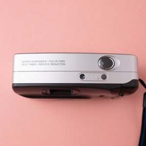 May include: A silver and black point-and-shoot camera with a black strap. The camera has the text "SUPER VIEWFINDER / FOCUS FREE SELF TIMER / RED-EYE REDUCTION" printed on the top.