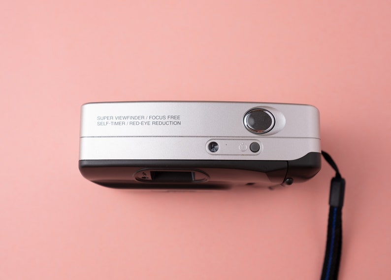May include: A silver and black point-and-shoot camera with a black strap. The camera has the text "SUPER VIEWFINDER / FOCUS FREE SELF TIMER / RED-EYE REDUCTION" printed on the top.