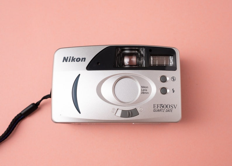 May include: A silver Nikon EF500SV Quartz Date camera with a black strap. The camera has a black lens with a 28mm lens.