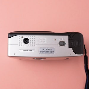 May include: A silver and black Nikon Coolpix camera with a black strap. The camera has a lens cover, a flash, and a zoom button. The camera is made in China and has a serial number of 6002361.