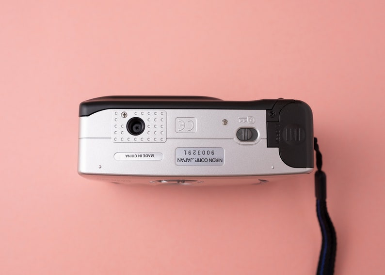 May include: A silver and black Nikon Coolpix camera with a black strap. The camera has a lens cover, a flash, and a zoom button. The camera is made in China and has a serial number of 6002361.