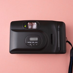 May include: A black film camera with a strap. The camera has a built-in flash and a small LCD screen with the text "MODE SEL ADJ".