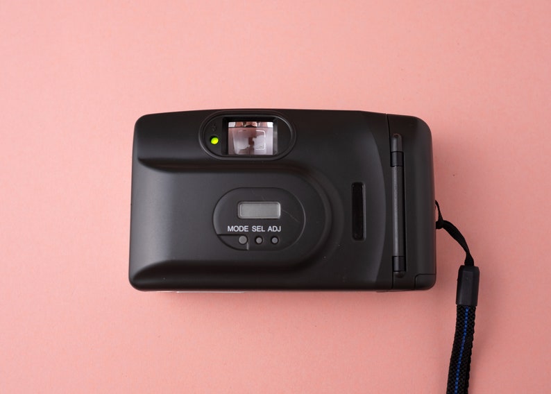 May include: A black film camera with a strap. The camera has a built-in flash and a small LCD screen with the text "MODE SEL ADJ".