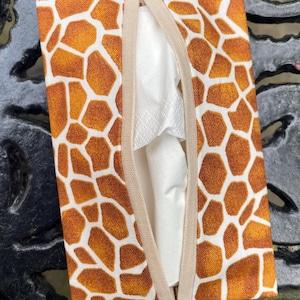 May include: A rectangular tissue box cover with a giraffe print in shades of brown and white. The cover has a beige elastic band on each side and a slit for tissue dispensing. White tissues are visible inside the cover.