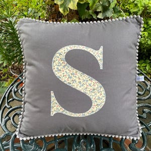 May include: A square grey throw cushion featuring a large, centred letter S appliquéd in a cream fabric with a delicate blue and pink floral pattern. The edges of the cushion are finished with a decorative grey pom-pom trim border.