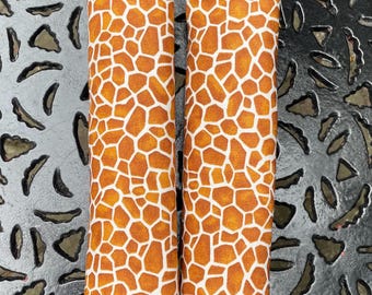 May include: Two fabric car seat belt covers with a giraffe print. The covers are rectangular and feature a pattern of brown and white irregular shapes. The background is a textured, metallic surface.