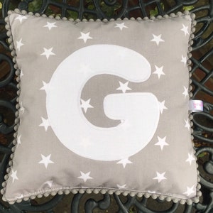 May include: A grey cushion with a white letter G on it. The cushion has a white star pattern and a grey pom-pom trim.