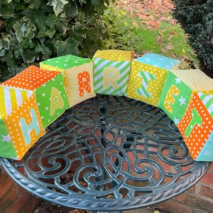 May include: A set of six colourful fabric blocks spelling out the name "HARRY". Each block is made of different patterned fabrics in shades of green, yellow, orange, and white. The blocks are arranged in a line on a black metal table.