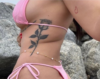 Pode incluir: Uma mulher vestindo um top e parte inferior de biquíni rosa com uma corrente dourada com pérolas brancas ao redor da cintura. Ela tem uma tatuagem de rosa preta no lado e uma pequena tatuagem de coração vermelho no braço.