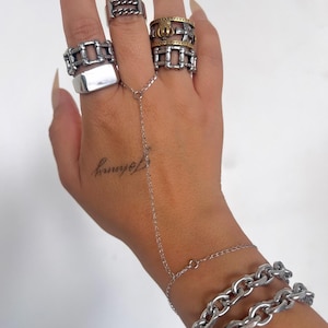Puede incluir: Una mano adornada con múltiples anillos plateados de varios diseños, incluyendo eslabones de cadena y formas rectangulares. Una cadena de mano plateada se conecta a una pulsera con un diseño de eslabones de cadena. La palabra "Johnny" está tatuada en la mano.