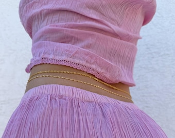 Könnte beinhalten: Ein Set aus einem hellrosa Oberteil und einem passenden Rock aus knittrigem Stoff. Das Oberteil hat einen zarten Spitzenbesatz am unteren Saum. Ein dünner, zweilagiger goldfarbener Ketten-Gürtel liegt um die Taille und betont den plissierten Stoff.