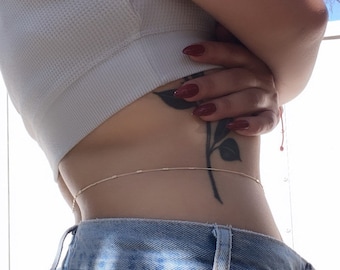 Könnte beinhalten: Eine Frau trägt ein weißes Tanktop und eine blaue Jeans mit einer zarten goldenen Kette um die Taille. Sie hat ein Blumen-Tattoo an der Seite und roten Nagellack.