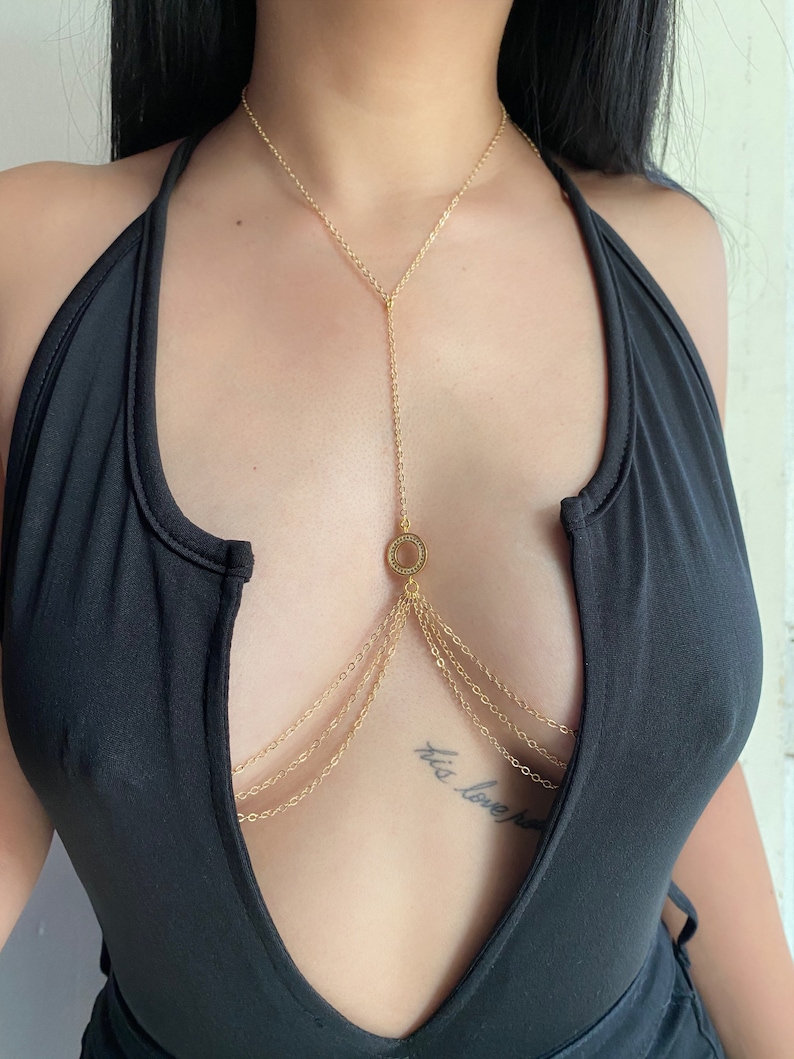 May include: A gold body chain with multiple layers of chains and a circular pendant. The chain is worn over a black top.