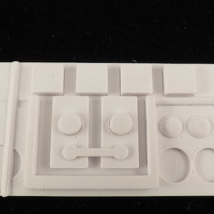 Può includere: Pannello in plastica bianca con una serie di pulsanti, manopole e interruttori. Il pannello ha una superficie testurizzata e sembra essere parte di un modello o di un giocattolo.