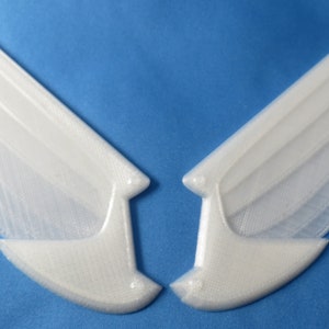 May include: Two white, 3D-printed angel wings, each with multiple layered sections, are displayed against a blue background. The wings are symmetrical, with a smooth, curved design, and are likely for crafting or costume use.