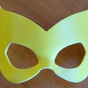 May include: A bright yellow eye mask featuring a pointed, cat-like silhouette with two distinct eye cutouts. The mask has a smooth, solid surface and a symmetrical design that tapers to sharp points at the top outer edges and rounded bottom.