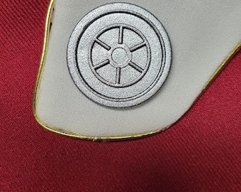 Könnte beinhalten: Ein silberfarbenes, kreisförmiges Emblem mit einem stilisierten Radmotiv, auf einem weißen Hintergrund mit goldener Umrandung. Das Emblem ist auf einer roten Stoffoberfläche befestigt.