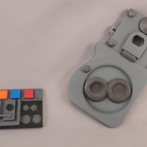 Op de afbeelding: Een grijs kunststof bedieningspaneel met drie knoppen, waarvan twee blauw en één oranje. Het paneel heeft een zwarte achtergrond met een grijs ontwerp. Een grijs kunststof bedieningspaneel met twee ronde knoppen en een hendel. Het paneel heeft een zwarte achtergrond met een grijs ontwerp.