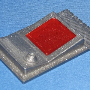 May include: A rectangular, silver-colored object with a raised border and a central, red, diamond-shaped insert. The object has a textured surface and a small, circular detail on one corner.