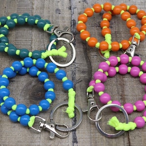 Könnte beinhalten: Vier Schlüsselanhänger mit Perlenarmband in Grün, Orange, Blau und Pink. Jeder Schlüsselanhänger hat runde Perlen, die mit einer neongelben Kordel aufgefädelt sind. Die Schlüsselanhänger haben einen silberfarbenen Verschluss und einen Schlüsselring.