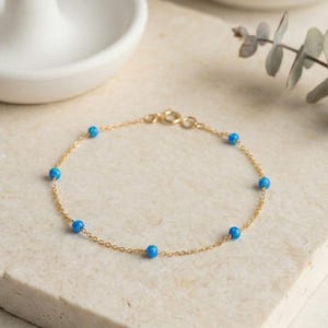 May include: A delicate gold chain bracelet featuring small, round, bright blue beads evenly spaced along the chain. The bracelet is displayed on a light-colored, textured surface, with a white ceramic dish and greenery in the background.