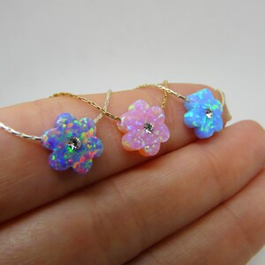 Puede incluir: Tres collares con colgantes de &oacute;palo en forma de flor en azul, rosa y celeste. Cada colgante tiene una peque&ntilde;a piedra central brillante y est&aacute; ensartado en una delicada cadena. Los collares se muestran sobre un fondo claro.