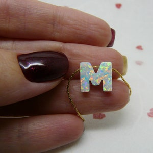 May include: A gold chain necklace with a white opal letter M pendant. The pendant has a rainbow iridescent effect.