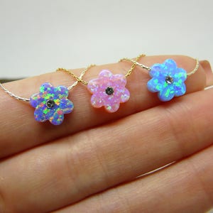 Puede incluir: Tres collares con colgantes en forma de flor de &oacute;palo iridiscente. Los colgantes son en tonos azul, rosa y azul-verdoso, cada uno con una peque&ntilde;a piedra oscura en el centro. Los collares se muestran en una mano.