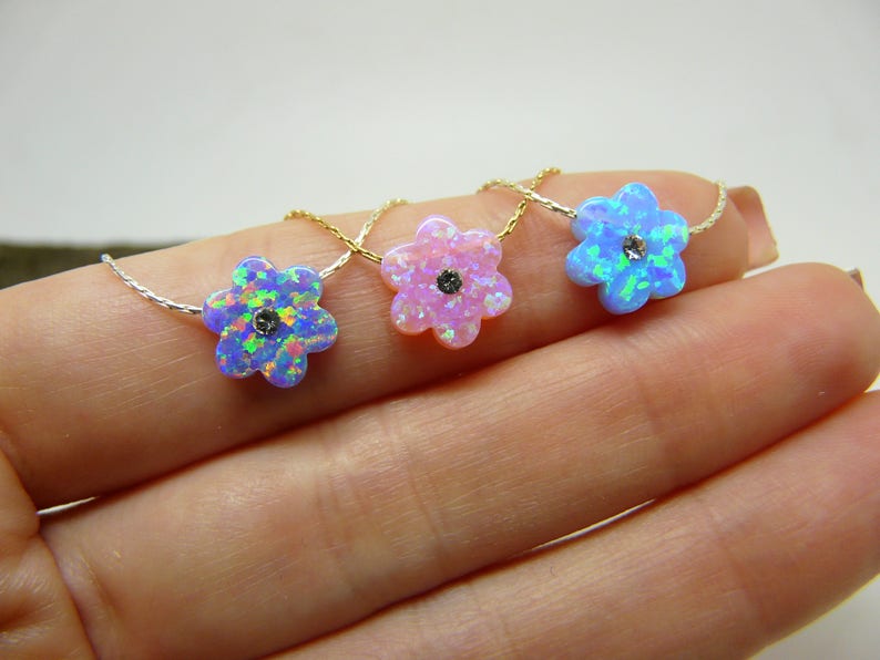 Puede incluir: Tres collares con colgantes en forma de flor de &oacute;palo iridiscente. Los colgantes son en tonos azul, rosa y azul-verdoso, cada uno con una peque&ntilde;a piedra oscura en el centro. Los collares se muestran en una mano.