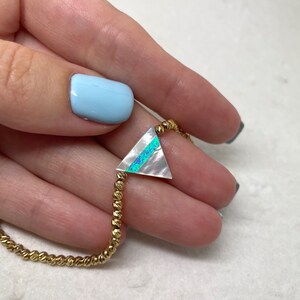 May include: A gold beaded bracelet with a triangular pendant. The pendant has a white mother-of-pearl base with a diagonal stripe of blue and green iridescent material. The bracelet is held in a hand with light blue nail polish.