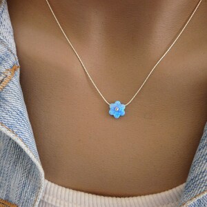 Puede incluir: Un delicado collar de plata con un colgante de flor azul claro con una peque&ntilde;a piedra central brillante. El colgante de la flor tiene seis p&eacute;talos. El collar se lleva sobre una chaqueta vaquera.