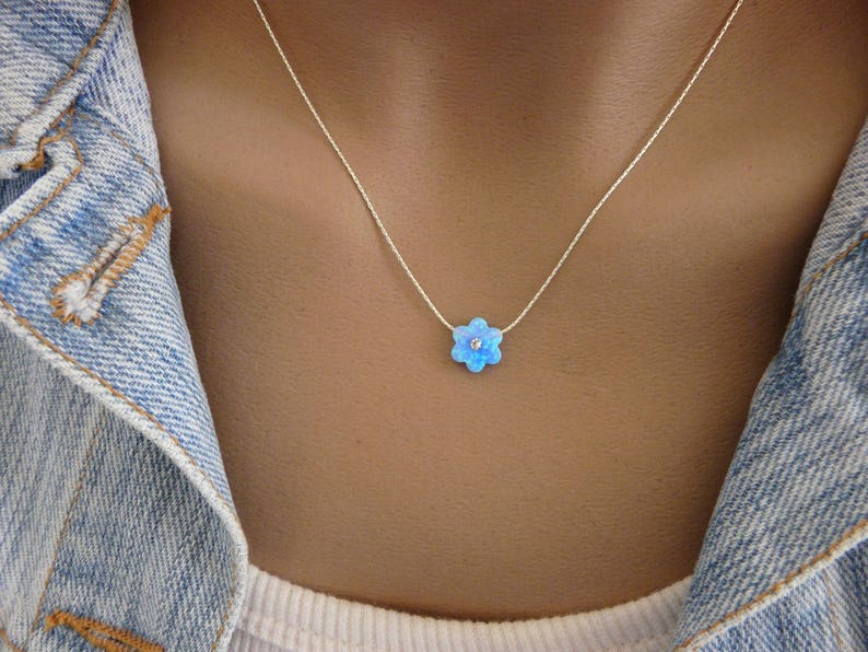 Puede incluir: Un delicado collar de plata con un colgante de flor azul claro con una peque&ntilde;a piedra central brillante. El colgante de la flor tiene seis p&eacute;talos. El collar se lleva sobre una chaqueta vaquera.