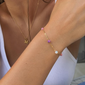 May include: A delicate gold chain bracelet featuring small, evenly spaced round beads in shades of pink, purple, and iridescent white. The bracelet is worn on a wrist, and a gold necklace with a letter M pendant is visible in the background.