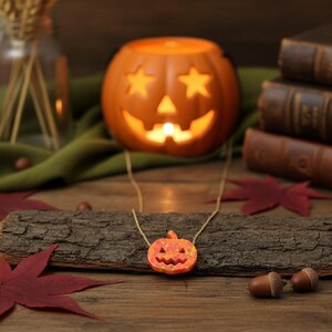 Puede incluir: Un collar de cadena dorada con un peque&ntilde;o colgante de calabaza naranja. La calabaza tiene rasgos faciales tallados. El collar se exhibe sobre un trozo de madera con hojas rojas y bellotas. Un candelabro de calabaza iluminado est&aacute; en el fondo.