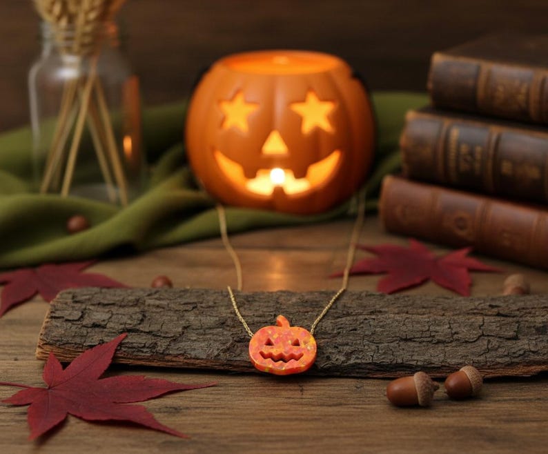 Puede incluir: Un collar de cadena dorada con un peque&ntilde;o colgante de calabaza naranja. La calabaza tiene rasgos faciales tallados. El collar se exhibe sobre un trozo de madera con hojas rojas y bellotas. Un candelabro de calabaza iluminado est&aacute; en el fondo.