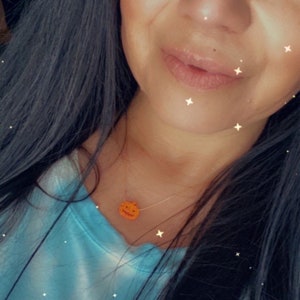 Puede incluir: Un collar dorado con un peque&ntilde;o colgante de calabaza naranja con rasgos faciales tallados. La calabaza est&aacute; suspendida de una delicada cadena. La persona lleva una camiseta azul claro.