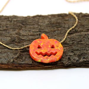 Puede incluir: Un collar tem&aacute;tico de Halloween con un colgante de calabaza tallada en tonos naranja y amarillo. El colgante est&aacute; suspendido de una delicada cadena dorada. El collar se exhibe sobre una superficie de madera marr&oacute;n texturizada.