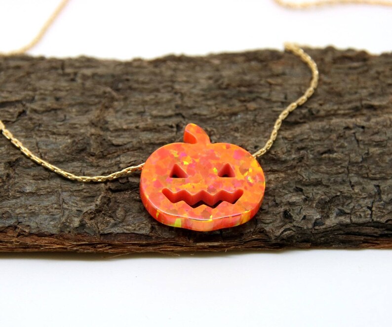 Puede incluir: Un collar tem&aacute;tico de Halloween con un colgante de calabaza tallada en tonos naranja y amarillo. El colgante est&aacute; suspendido de una delicada cadena dorada. El collar se exhibe sobre una superficie de madera marr&oacute;n texturizada.