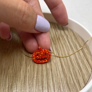 Puede incluir: Un collar dorado con un peque&ntilde;o colgante de calabaza naranja. La calabaza tiene un dise&ntilde;o de cara tallada y est&aacute; engastada con piedras naranjas brillantes. El collar se muestra sobre una superficie de madera.