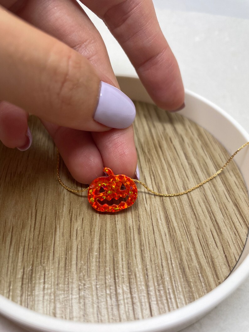 Puede incluir: Un collar dorado con un peque&ntilde;o colgante de calabaza naranja. La calabaza tiene un dise&ntilde;o de cara tallada y est&aacute; engastada con piedras naranjas brillantes. El collar se muestra sobre una superficie de madera.