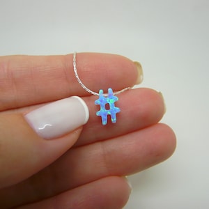 May include: A silver necklace with a blue opal hashtag pendant. The pendant is light blue with green accents. The necklace is held in a hand against a white backdrop.