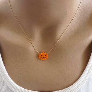 Puede incluir: Un collar con cadena dorada y un peque&ntilde;o colgante de calabaza naranja. La calabaza tiene un dise&ntilde;o de cara tallada, perfecto para Halloween. El collar se lleva alrededor del cuello.