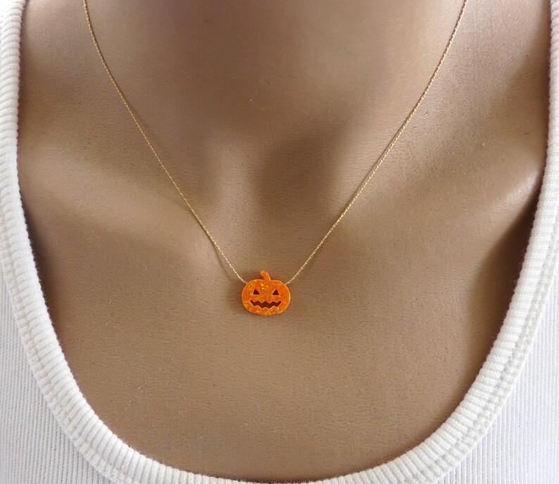 Puede incluir: Un collar con cadena dorada y un peque&ntilde;o colgante de calabaza naranja. La calabaza tiene un dise&ntilde;o de cara tallada, perfecto para Halloween. El collar se lleva alrededor del cuello.
