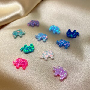 May include: Assortment of small, iridescent opal elephant figurines in various colors, including pink, blue, green, and white. The figurines are arranged on a light beige fabric surface.