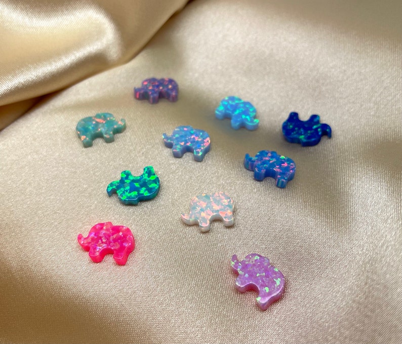 May include: Assortment of small, iridescent opal elephant figurines in various colors, including pink, blue, green, and white. The figurines are arranged on a light beige fabric surface.
