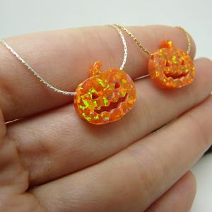 Puede incluir: Dos collares con colgantes de calabaza naranja. Los colgantes tienen caras talladas y est&aacute;n llenos de acentos coloridos y brillantes. Los collares se muestran sobre cadenas plateadas y doradas, sobre un fondo claro.