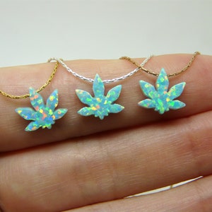 May include: Three opalite marijuana leaf pendants on delicate gold and silver chains. The pendants are light blue with iridescent flecks, and the chains are held by a person's hand.