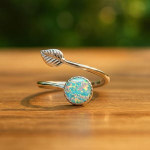 May include: A silver-toned ring with a leaf design and a round, opalescent stone. The ring features a leaf-shaped accent on one end and a bezel-set, blue-green opal at the other. The ring is set against a blurred green background.