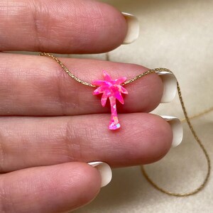 以下が含まれることがあります： 繊細なゴールドチェーンネックレスに、ピンクオパールのヤシの木のペンダントが飾られています。ペンダントは、キラキラと輝くピンクの仕上げが施された、小さな手の込んだヤシの木です。