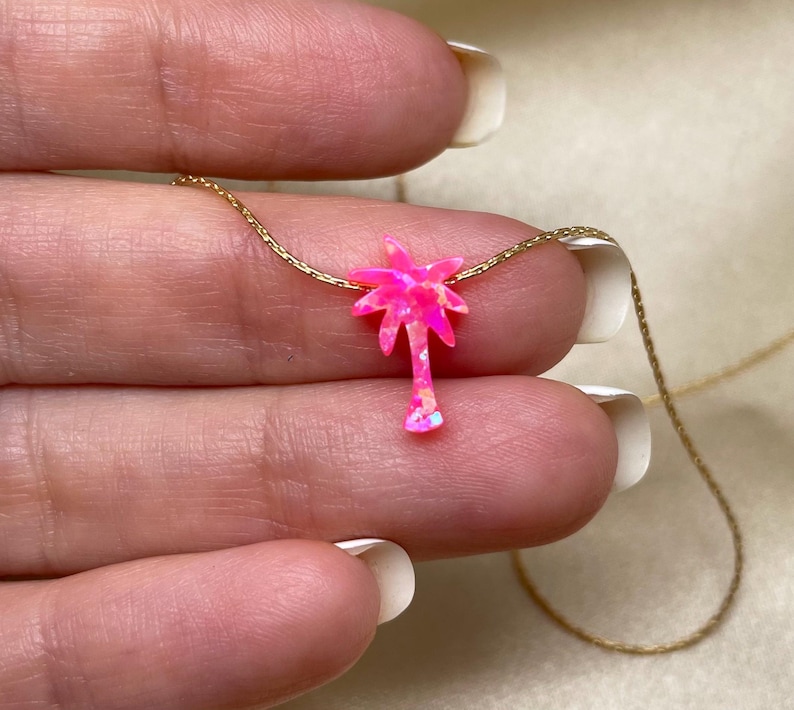 以下が含まれることがあります： 繊細なゴールドチェーンネックレスに、ピンクオパールのヤシの木のペンダントが飾られています。ペンダントは、キラキラと輝くピンクの仕上げが施された、小さな手の込んだヤシの木です。